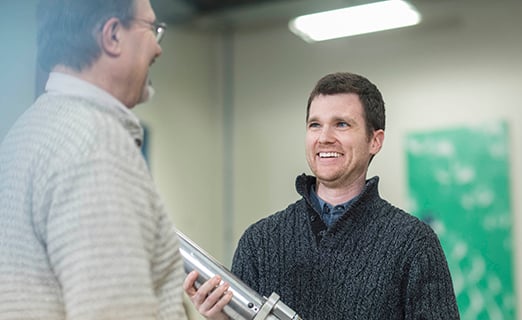 Person is discussing with a colleague while holding a machine part.